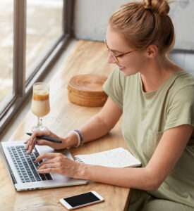 Smiling woman with glasses and a hair bun typing on a laptop at a wooden cafe counter next to a window, with an open notebook, pen, and glass of iced coffee nearby.