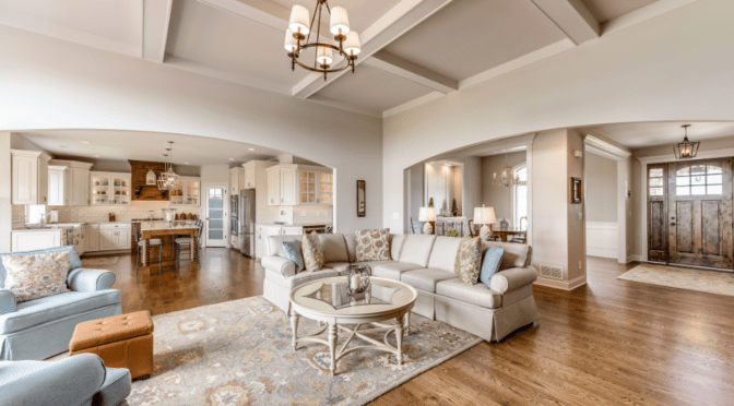 Open-concept living space with a cozy living room, elegant kitchen, and welcoming entryway. The living room features a large beige sectional sofa, patterned throw pillows, and a round glass coffee table atop a decorative area rug. Adjacent is a spacious kitchen with white cabinetry, a wooden island with bar stools, and pendant lighting.