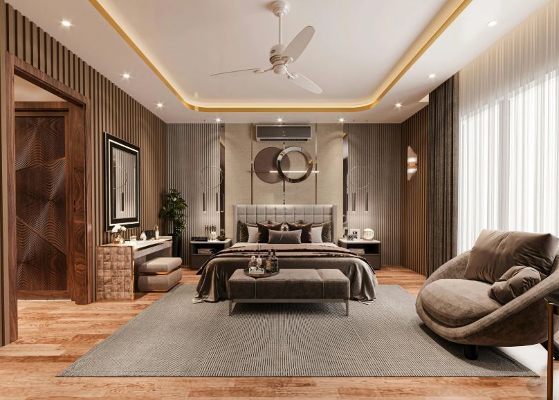A modern luxury bedroom interior featuring a grey upholstered bed with brown bedding, fluted wooden accent walls, recessed ceiling lighting with gold trim, a ceiling fan, a large grey rug, a sculptural armchair, and decorative circular wall art above the bed.