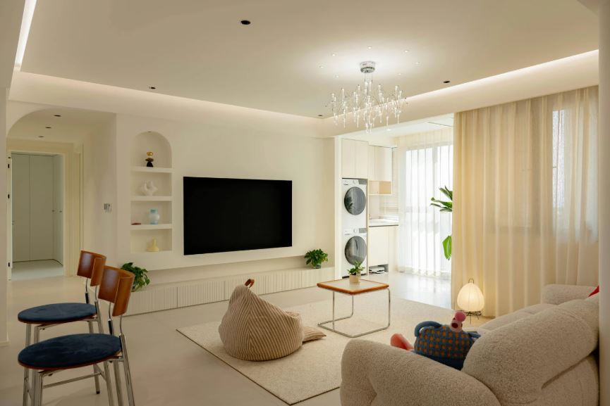 A bright, modern living room with a neutral colour palette, featuring a wall-mounted TV with built-in shelving, a cosy sofa and bean bag seating, soft sheer curtains filtering daylight, and minimalist furniture accented with indoor plants.