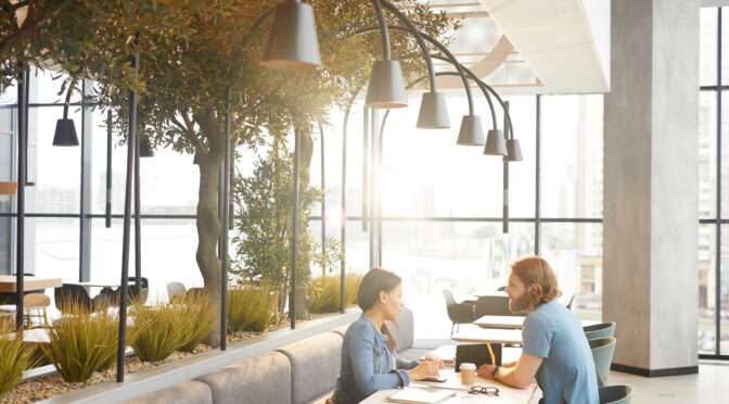 Two people having a meeting in a modern biophilic-designed office café with natural lighting, indoor plants, and contemporary furnishings.
