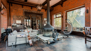 An eclectic industrial living room with exposed brick walls, a suspended metal fireplace, mixed vintage furniture, large windows, and artistic décor elements.