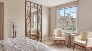 A calm bedroom corner with two upholstered armchairs by a large window, a decorative floor mirror with a geometric frame, soft neutral walls, and a cityscape view outside.