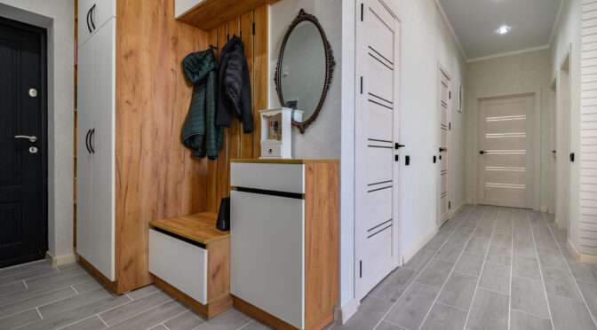 Modern hallway with wooden and white storage cabinets, wall-mounted mirror, coat hooks, and gray tile flooring.