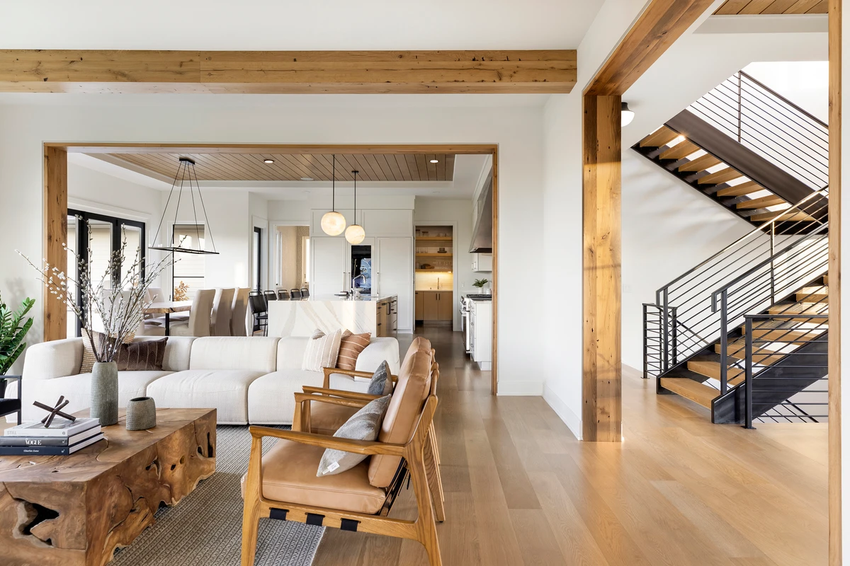 Modern open-plan living area with exposed timber beams, wooden staircase, neutral furnishings, and abundant natural light.
