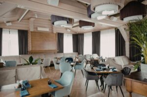 Modern restaurant dining area with soft upholstered seating, natural wood finishes, indoor plants, and warm ambient lighting.