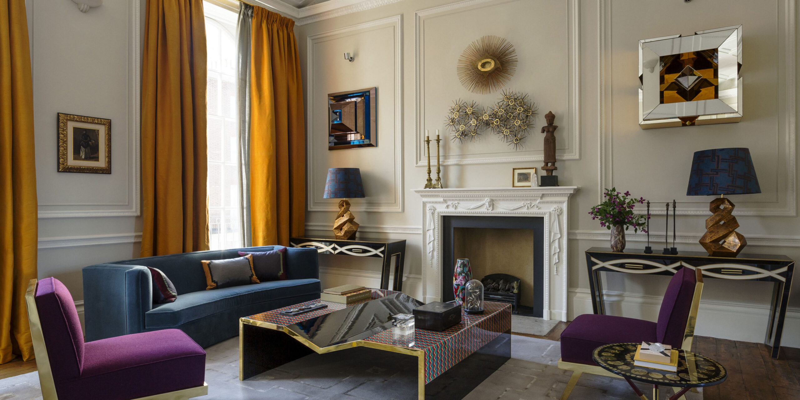 Elegant living room with classic fireplace, velvet seating, glass coffee table, decorative artwork, and warm accent colours in a refined interior.