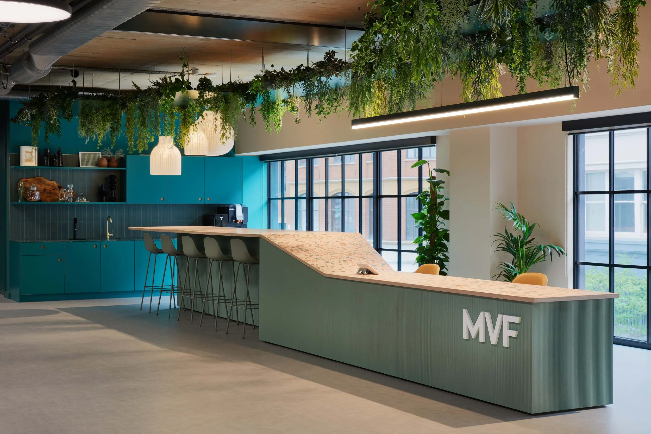 Modern office reception area with a green-toned desk, hanging plants, pendant lighting, and contemporary workspace design.