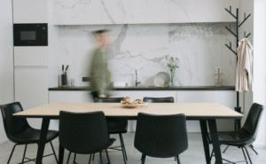 A minimalist kitchen–dining space with a light wood dining table, black dining chairs, white marble-effect cabinetry, open shelving, and a coat stand, with a blurred figure moving through the space.