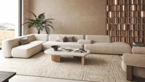 A minimalist living room with a low modular sectional sofa in soft neutral tones, a sculptural coffee table, textured rug, indoor plants, and warm textured wall finishes.