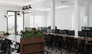 A bright open-plan office with rows of computer workstations, ergonomic chairs, white columns, integrated planters, pendant lighting, and large windows providing natural light.