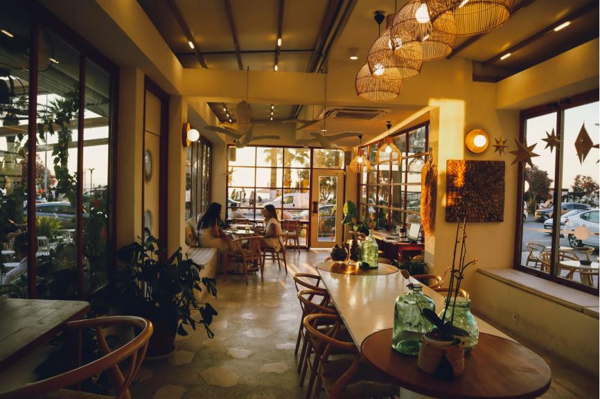 Cozy café interior with wooden tables and chairs, green glass vases, rattan pendant lights, large windows and potted plants.