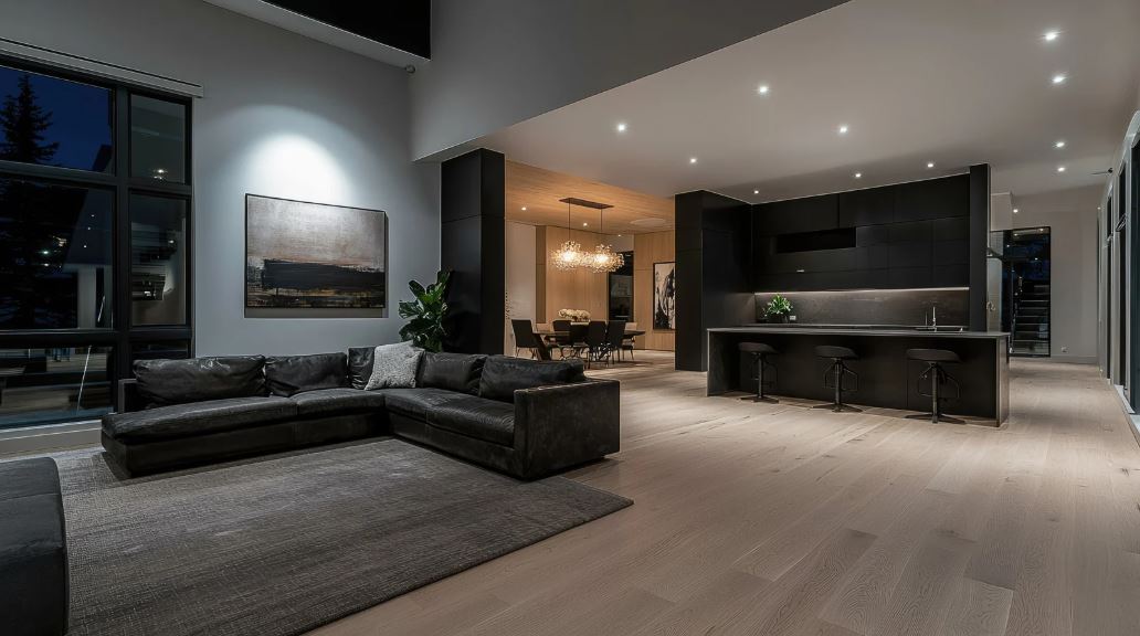 “Spacious open-plan living area with a low-profile sectional sofa, dark kitchen cabinetry, integrated lighting, floor-to-ceiling windows, and a sleek contemporary layout.”