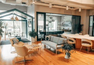 A contemporary office lounge with wooden flooring, casual seating, a sofa, armchairs, indoor plants, glass meeting rooms, and a bright open layout with natural light.
