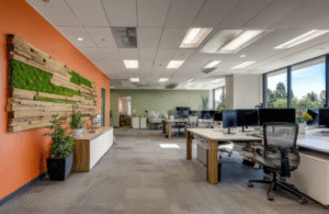 A modern open-plan office with workstations, task chairs, desktop monitors, a feature wall with greenery and timber accents, carpeted flooring, and large windows providing daylight.