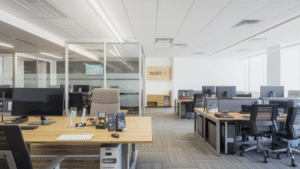 A clean, open-plan office with individual workstations, desktop monitors, ergonomic chairs, glass-partitioned rooms, neutral finishes, and evenly distributed ceiling lighting.