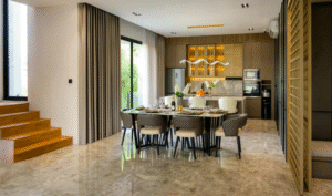 A modern dining area with a large dining table, upholstered chairs, pendant lighting, a contemporary kitchen in the background, polished stone flooring, and floor-to-ceiling curtains.