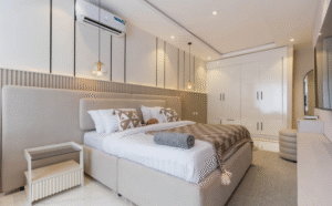 A modern minimalist bedroom with a low upholstered bed, layered neutral bedding, wall-mounted pendant lights, built-in wardrobes, soft ambient lighting, and a light monochrome colour palette.