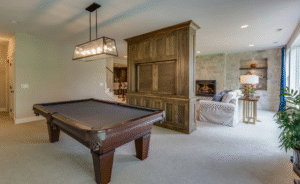A multi-purpose entertainment room featuring a pool table, a wooden built-in cabinet, pendant lighting, soft carpeted flooring, and a cosy seating area with a fireplace in the background.