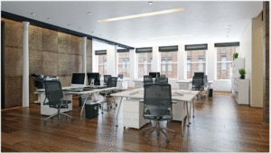 A contemporary open-plan office with shared desks, desktop computers, ergonomic mesh chairs, glass partitions, wood flooring, and large windows allowing natural light.