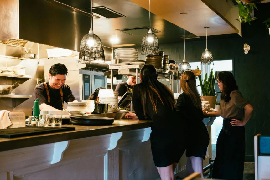 “Restaurant bar interior with guests seated at a counter, chefs working in an open kitchen, warm pendant lighting, and a modern, social dining atmosphere.”