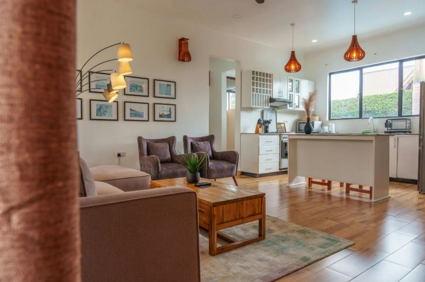 “Open-plan living and kitchen area with a comfortable sofa, wooden coffee table, soft rugs, pendant lighting, and a bright kitchen island with bar stools.”