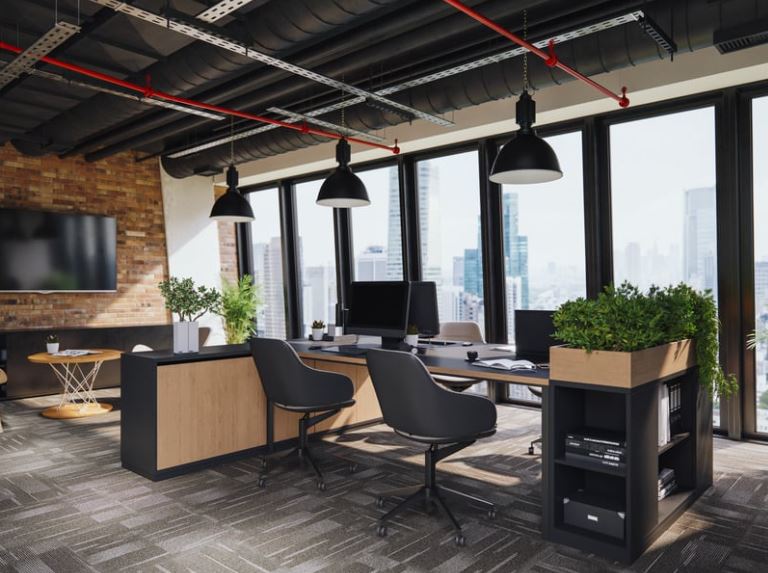 Modern office workspace with exposed black ceiling, red pipe, three pendant lights, brick wall, large windows, desk with black chair, and green plants.