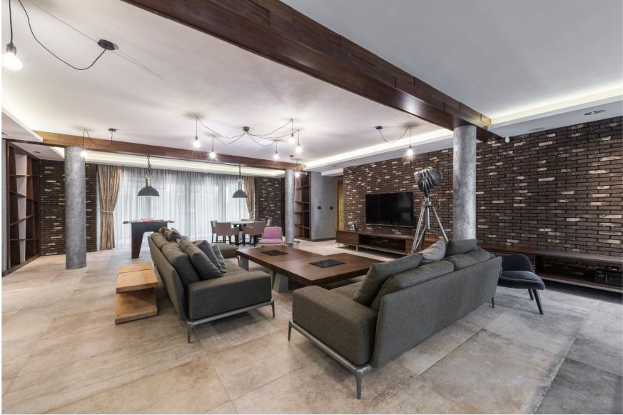 “Spacious contemporary living room with dark upholstered sofas, a large wooden coffee table, textured feature walls, stone flooring, and layered ambient lighting.”