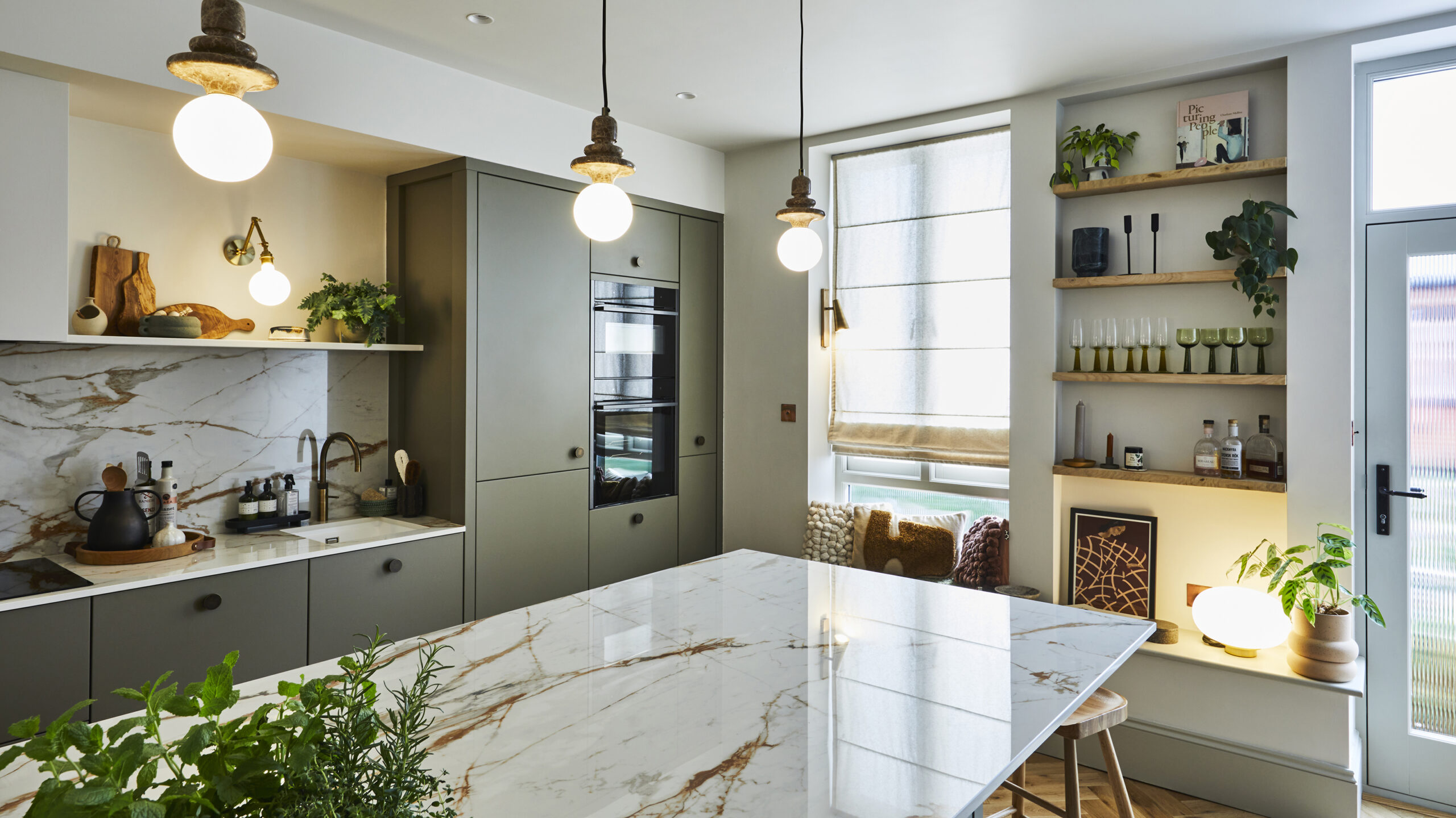 “Contemporary kitchen with sage green cabinetry, marble island worktop, open wooden shelving, pendant lighting, and large windows allowing natural daylight.”