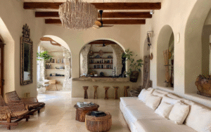 A Mediterranean-style living room with arched openings, exposed wooden ceiling beams, white plaster walls, a built-in bar with stools, woven furniture, and natural textures throughout.