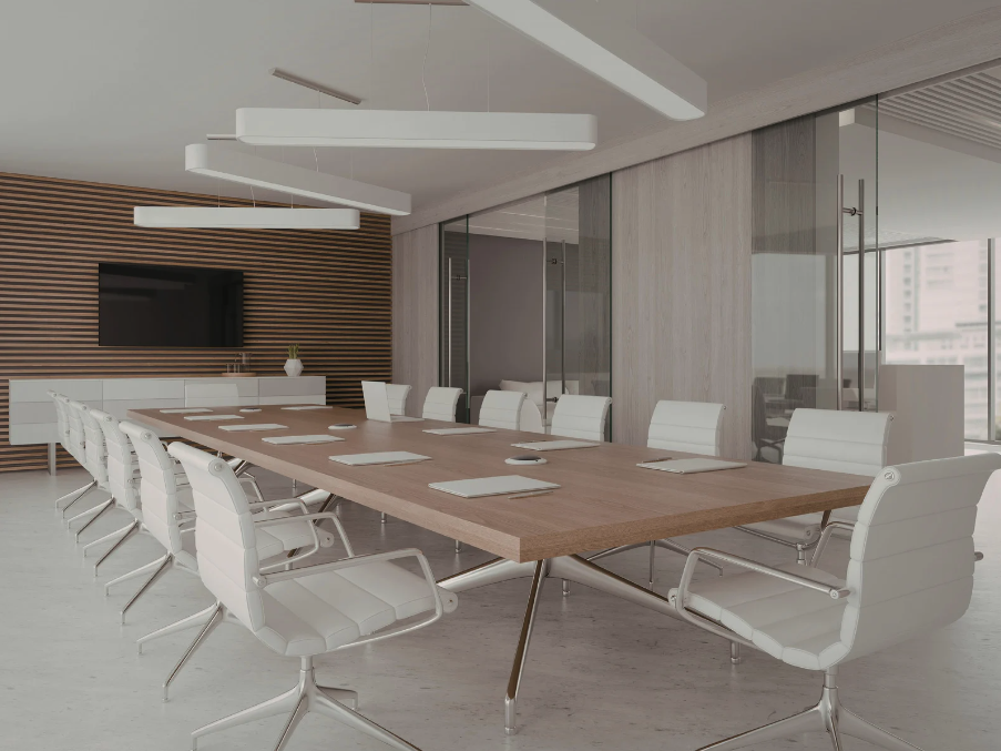 A modern corporate boardroom featuring a long wooden conference table, white ergonomic swivel chairs, minimalist pendant lights, a wall-mounted screen, and glass partitions with city views.