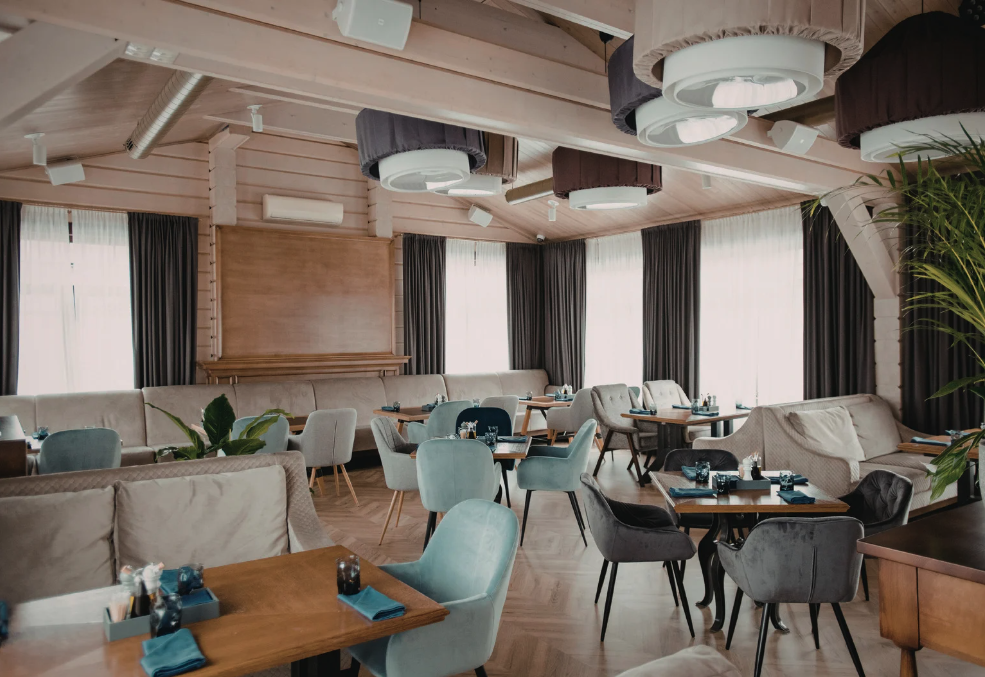 A cosy, modern restaurant interior with upholstered dining chairs, wooden tables, soft booth seating, exposed timber ceiling beams, fabric-wrapped ceiling lights, and large windows with dark curtains.