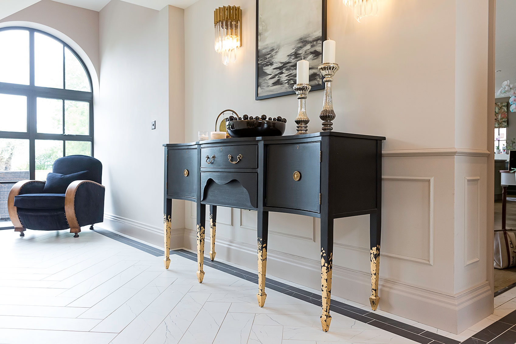 “Elegant hallway interior featuring a black console table with gold accents, decorative accessories, soft wall panelling, and natural light from an arched window.”