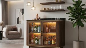 A wooden bar cabinet with glass doors, illuminated interior, black marble top, and brass accents, displaying bottles and glasses in a modern living room with floating shelves and a potted plant.