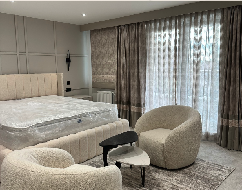 Contemporary Master Bedroom Design in London A soft neutral master bedroom featuring upholstered bed, lounge chairs, layered curtains, and textured wall panels, designed by Oraanj Interiors for a luxury London residence.