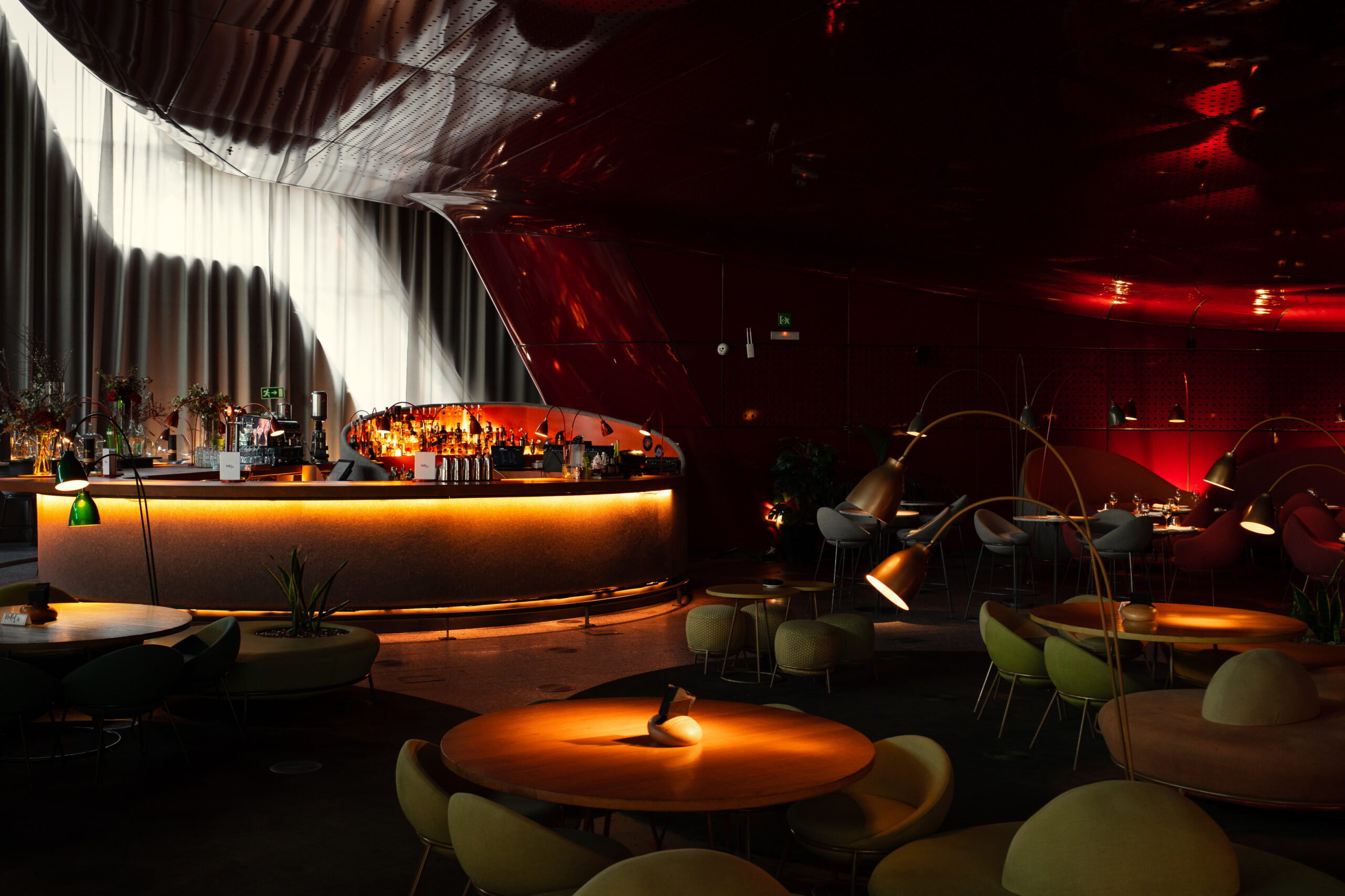 Moody lounge bar interior with a curved illuminated counter, red reflective ceiling, and round tables with modern seating.