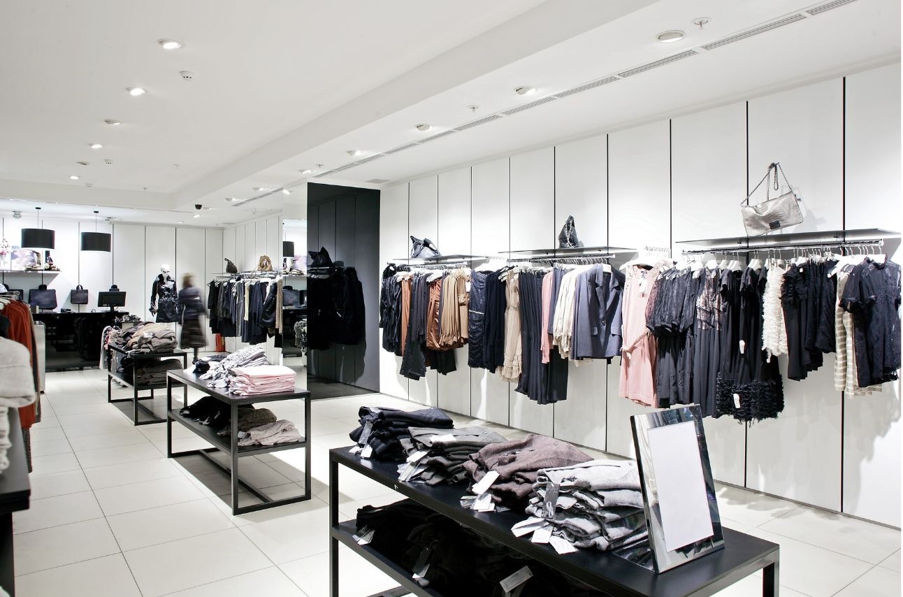 Modern retail clothing store interior with black display tables holding folded garments, wall racks of hanging clothes in neutral and black tones, white panelled walls and recessed lighting.