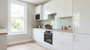 Bright tiny kitchen with glossy white cabinets, glass splashback, wooden countertops, brass handles, and natural light creating an airy spacious feel